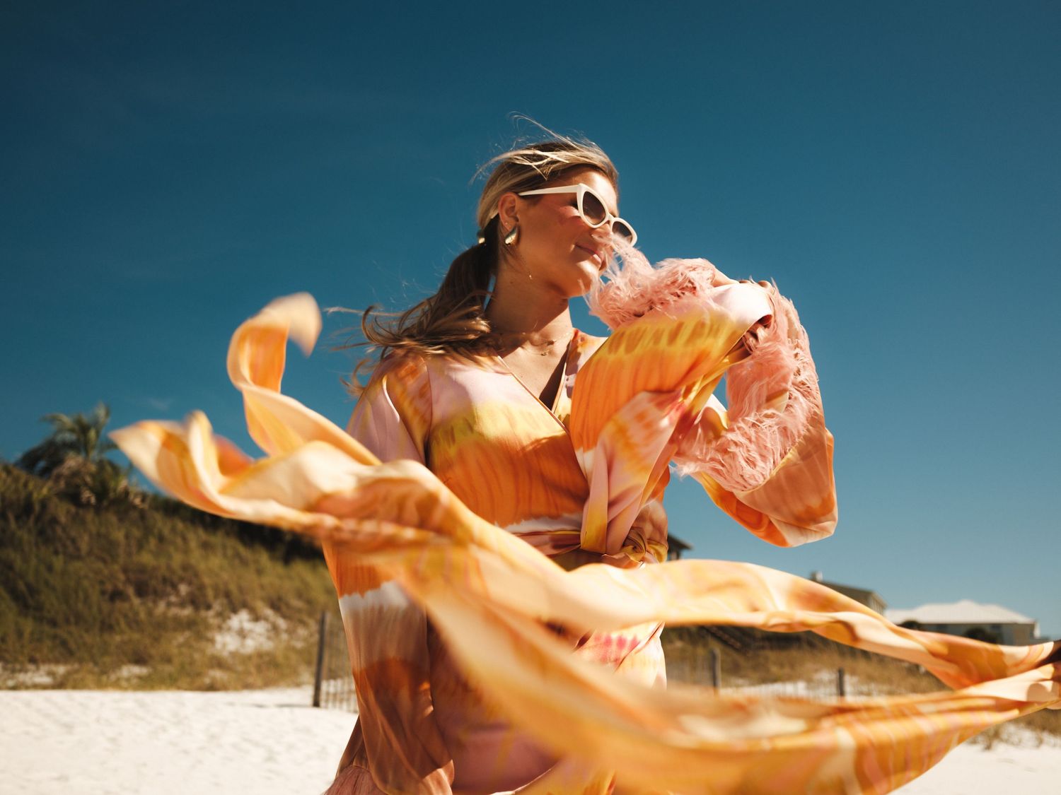 Woman in flowing orange boho dress dancing on a sunny beach with blue sky and sandy shores.