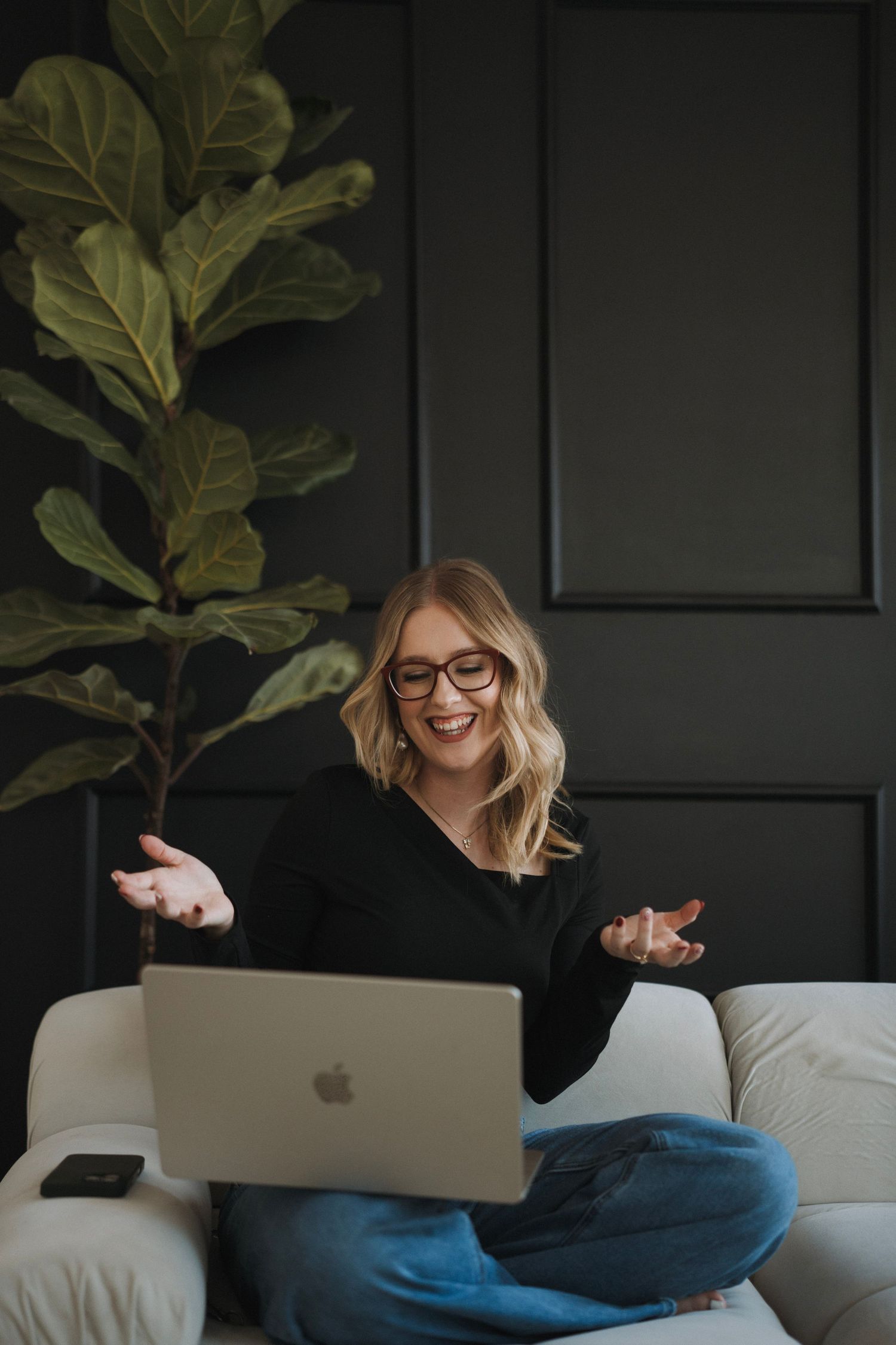 Smiling blonde woman with glasses using Apple MacBook laptop on white sofa next to fiddle leaf fig plant.