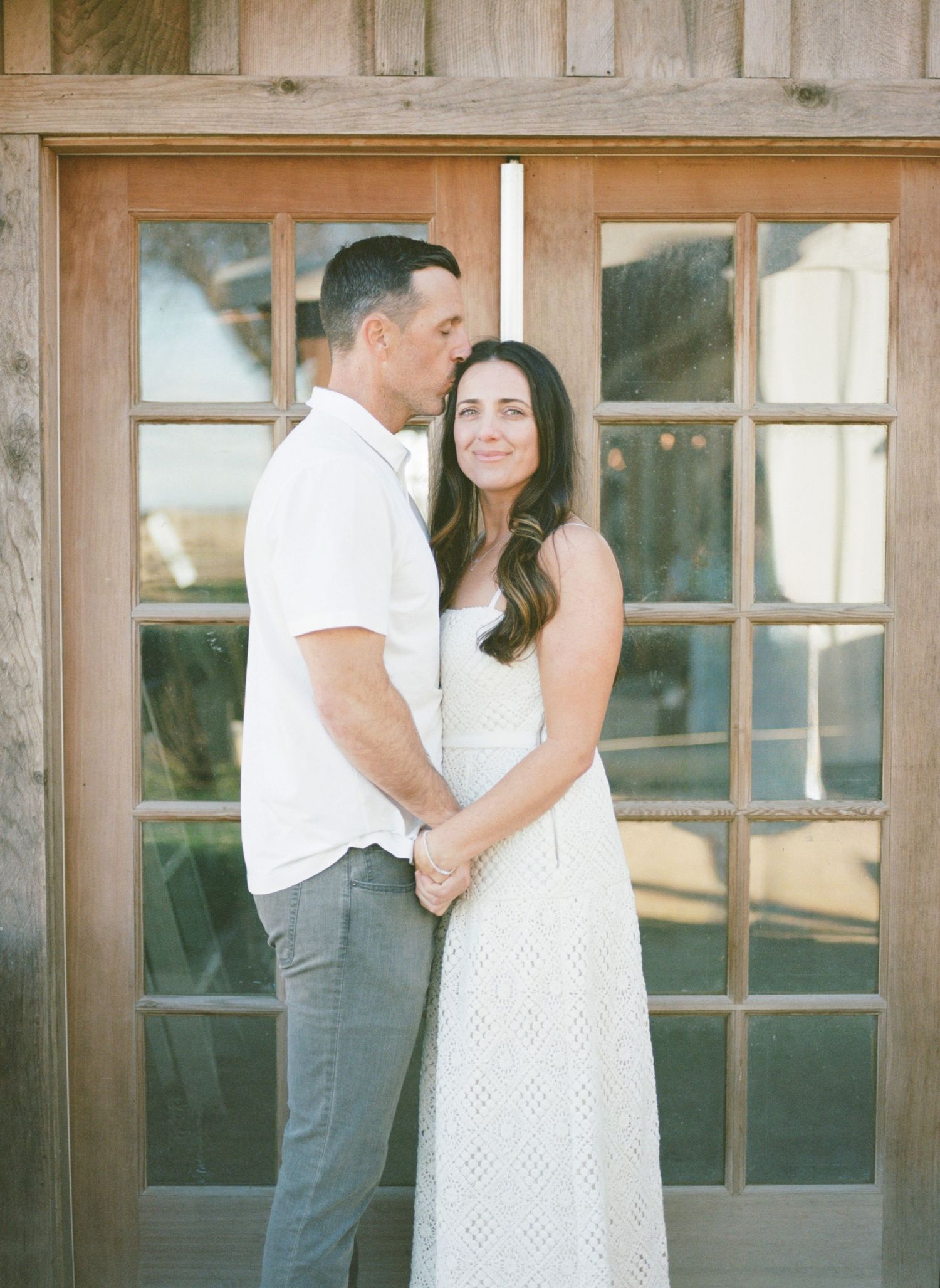 Couple holding hands in front of rustic wooden French doors, woman in white lace dress, man in white shirt and gray pants.