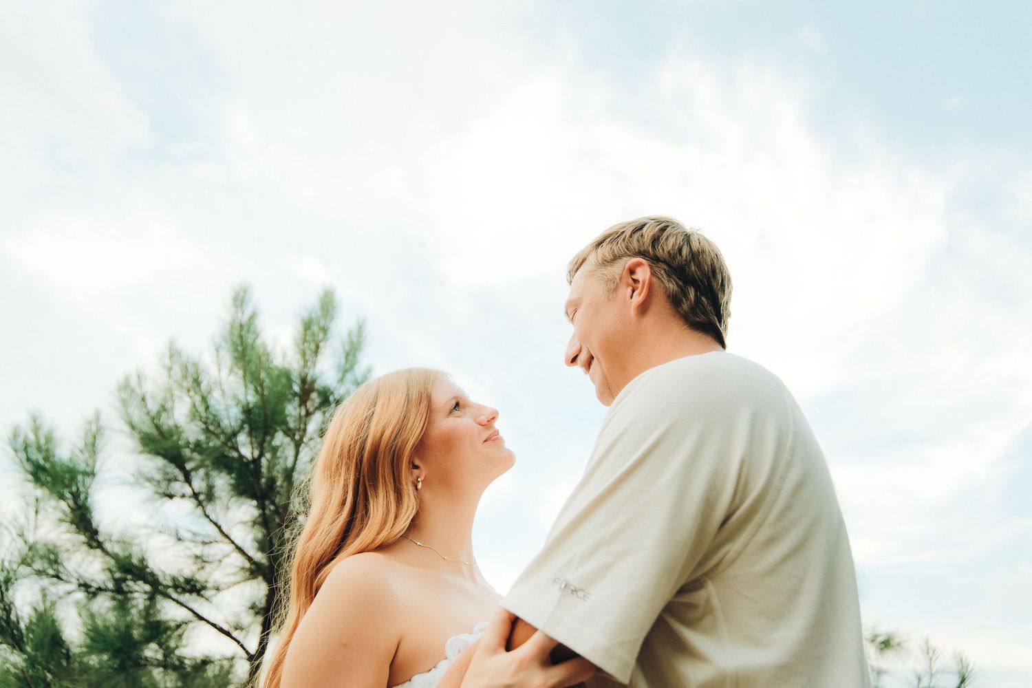Couple smiling at each other outdoors under a blue sky with pine trees in the background.
