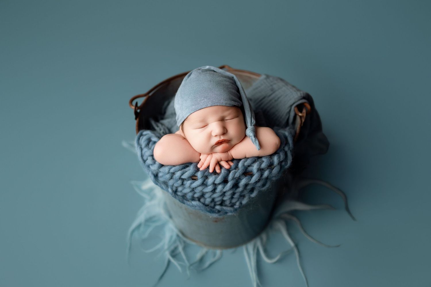 Sleeping newborn baby in blue knit bonnet nestled in a rustic metal bucket with chunky yarn blanket on teal background.