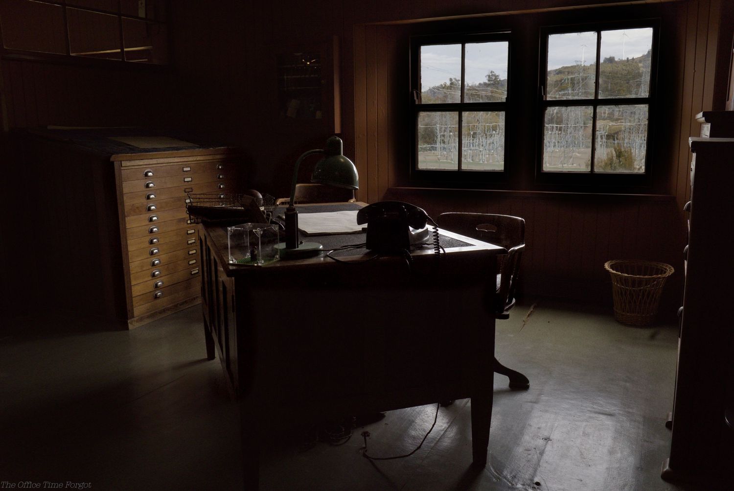 Dimly lit vintage office with a wooden desk, desk lamp, chair, and flat file cabinet beside two windows overlooking a green hillside.