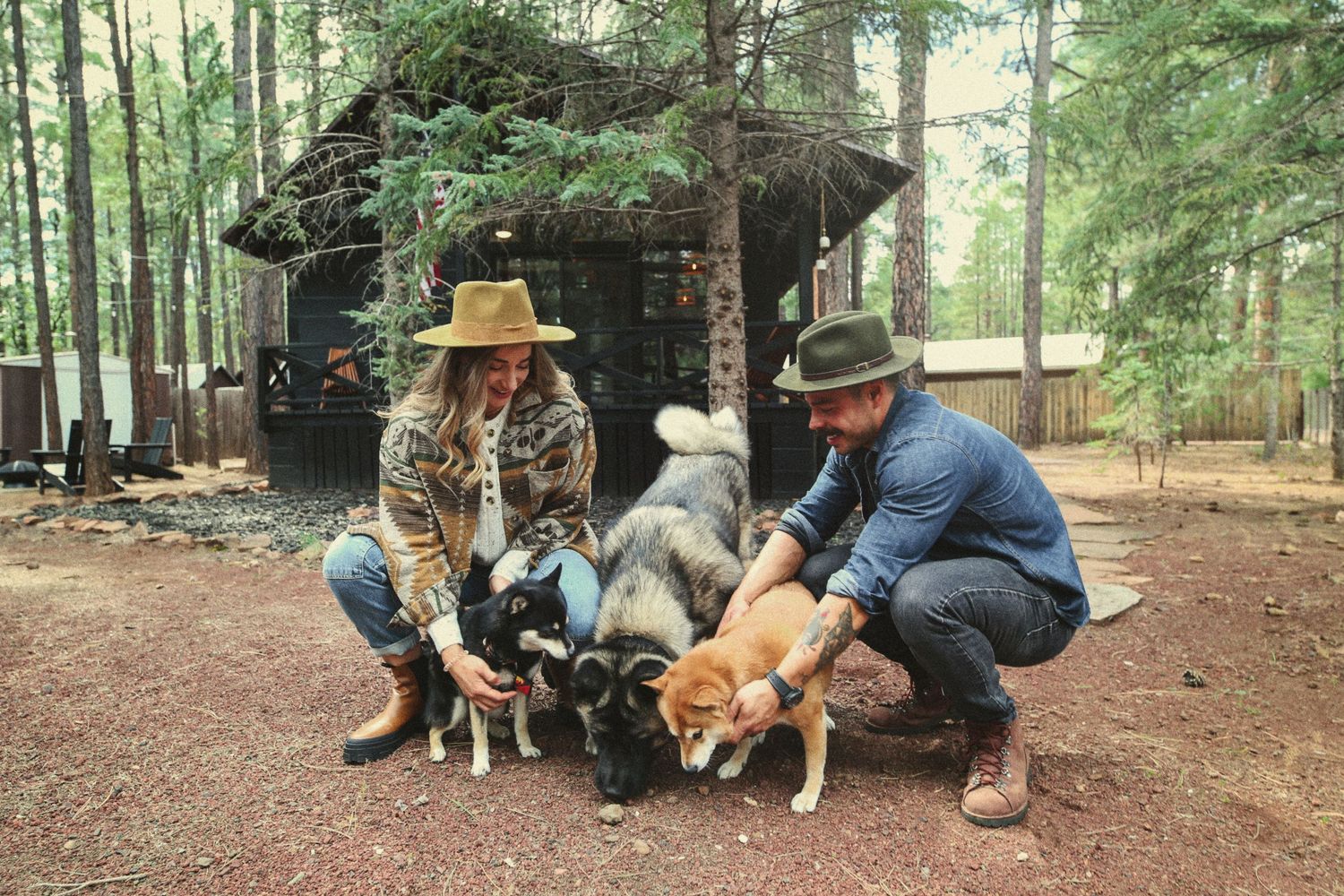 Two people in hats crouch on a dirt path in a pine forest, petting three dogs near a rustic wooden cabin.