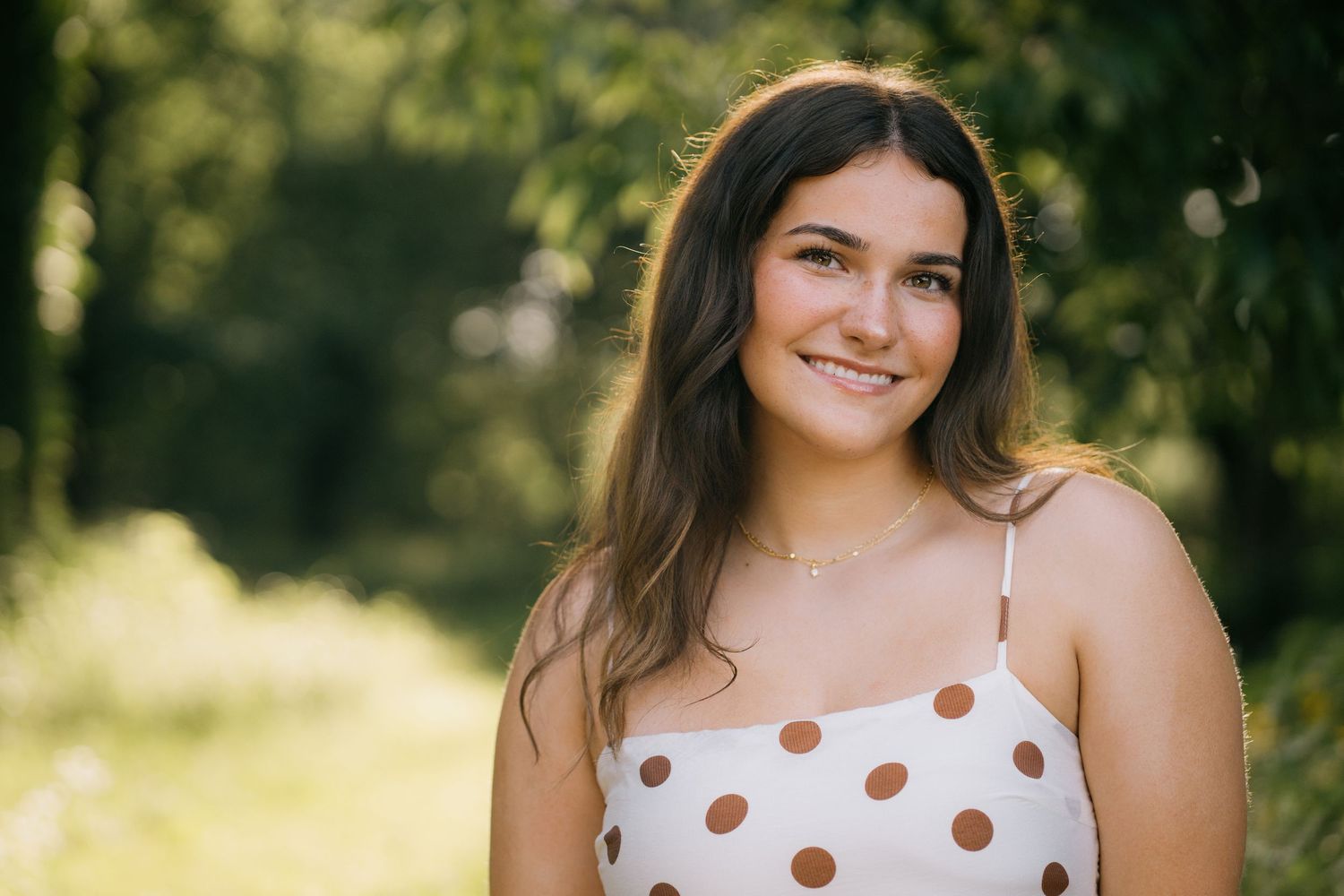 Smiling young woman with brown hair wearing a white and pink floral top, posing outdoors in natural light.