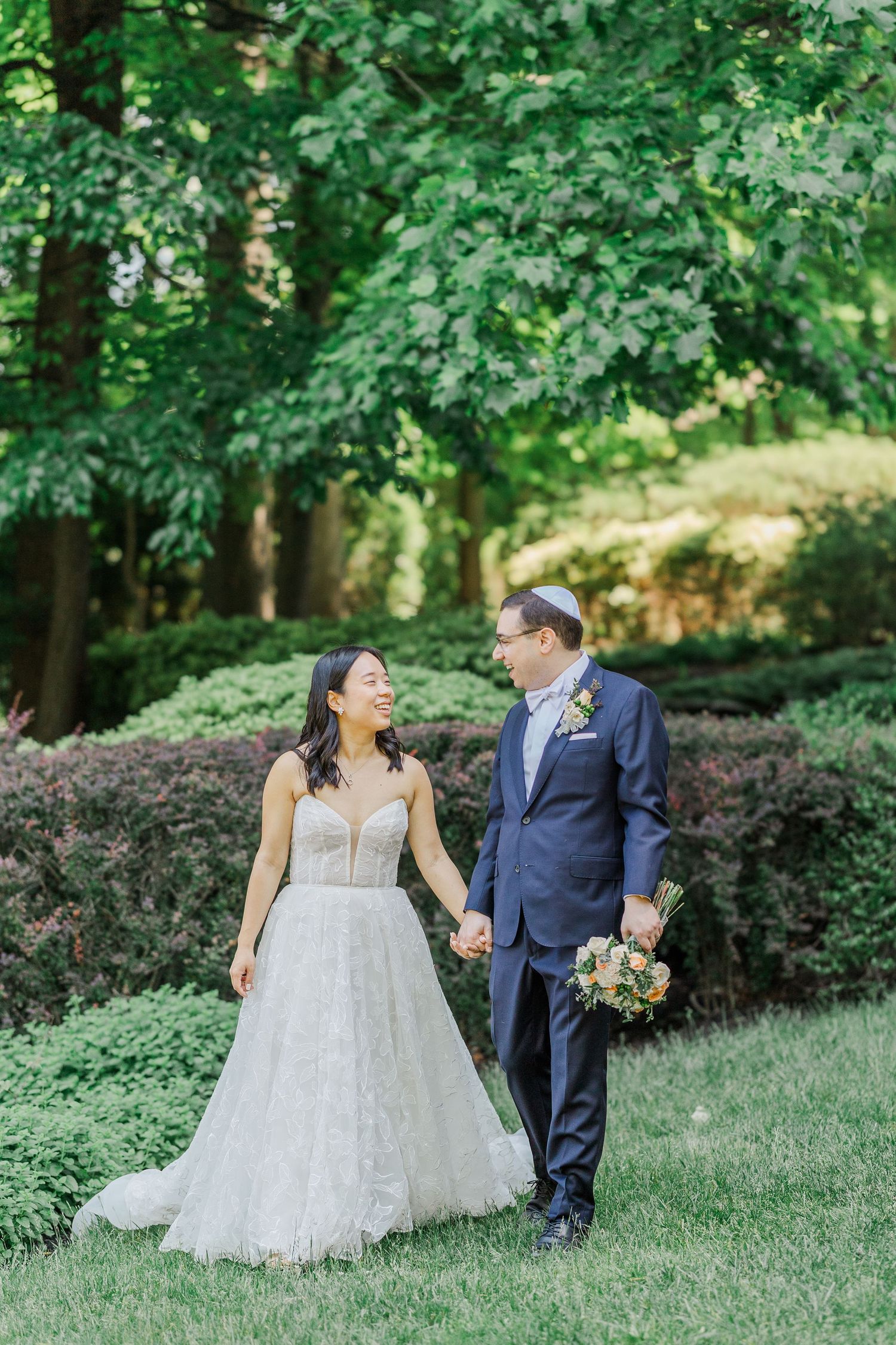 Bride and groom walk hand in hand through lush garden, she in white gown, he in navy suit with yarmulke.