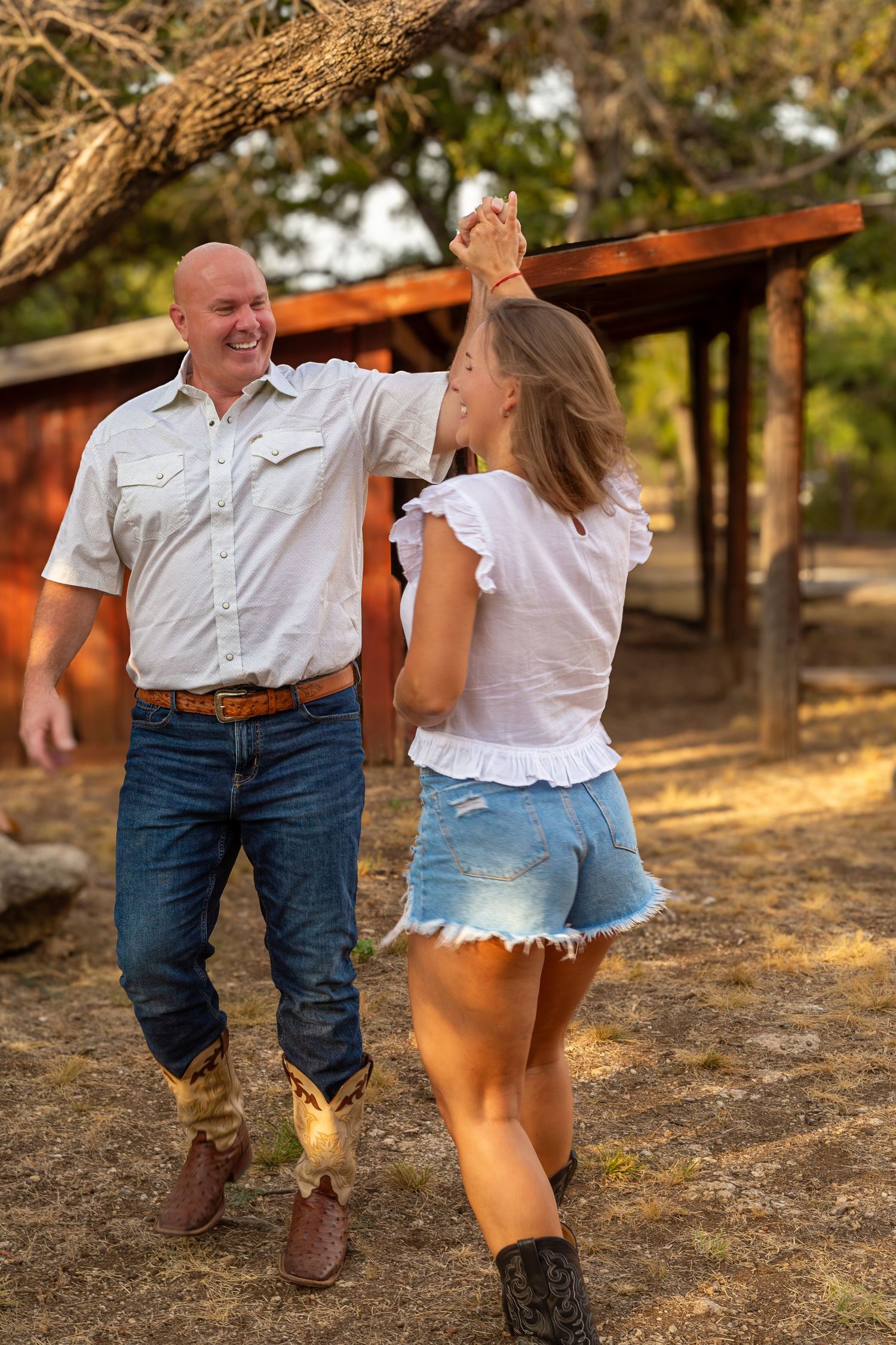 A bald man in jeans and boots dances with a young woman in denim shorts outdoors near a rustic wooden structure.