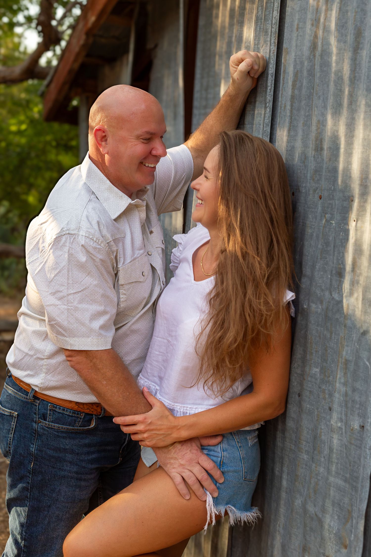 A smiling bald man in a white shirt leans against a rustic wall with a woman in denim shorts, sharing a romantic moment.