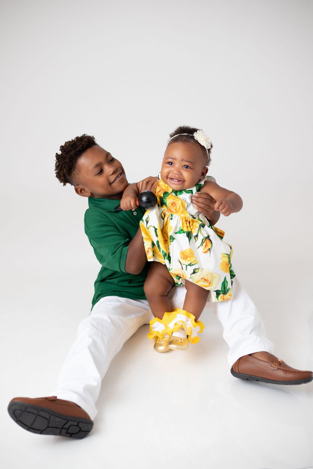 A young boy in a green shirt holds a smiling baby girl in a floral yellow dress on a white background.