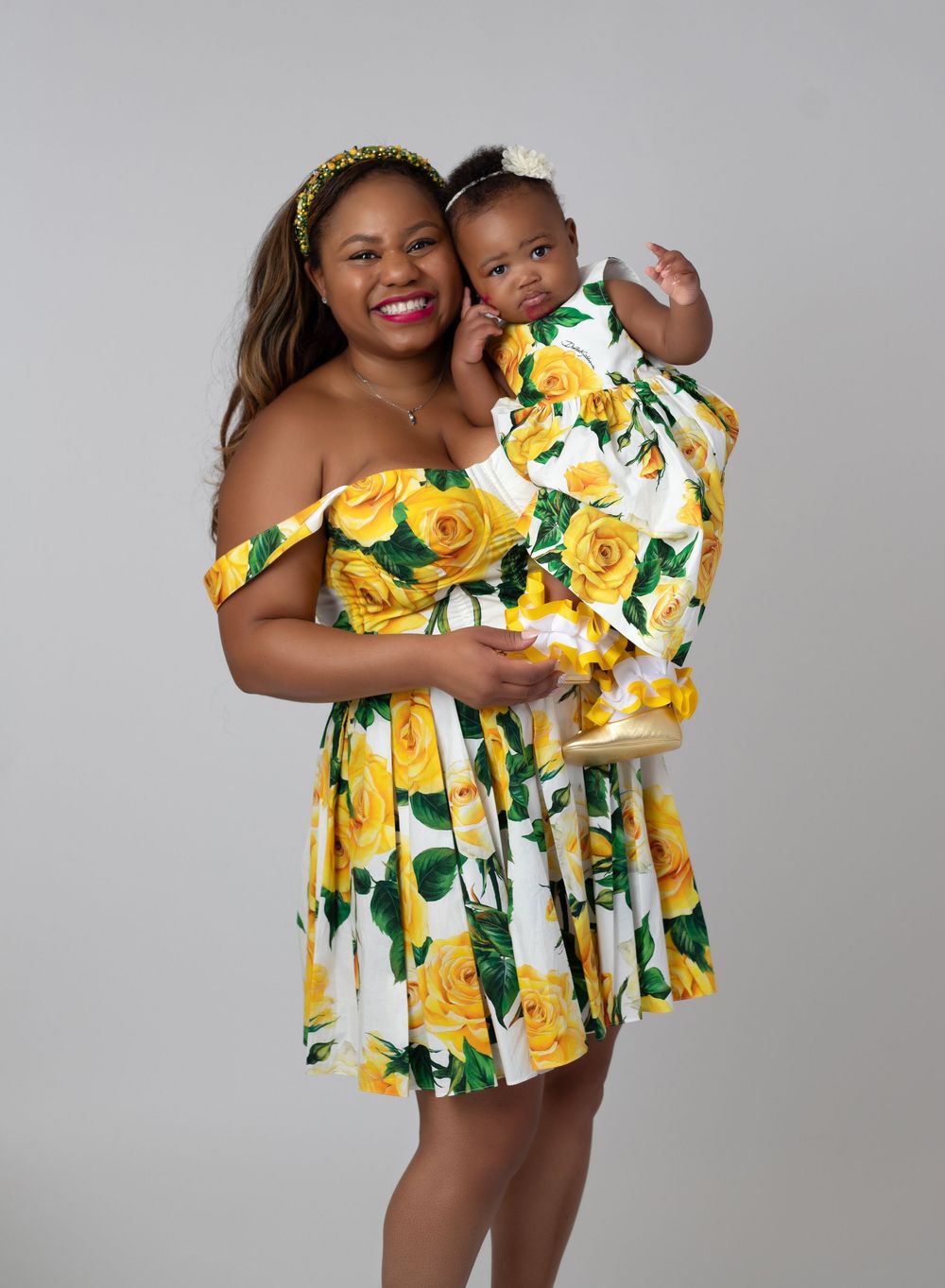 Mother and baby in matching yellow floral off-shoulder dresses, smiling together against a gray background.