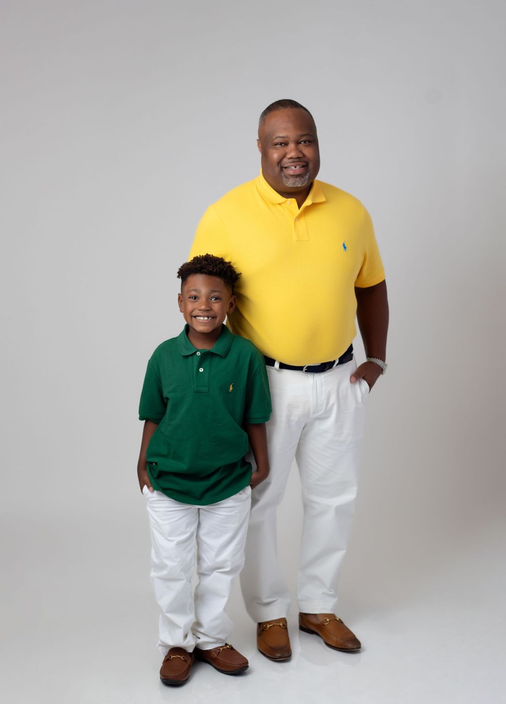 A man in a yellow polo and boy in green polo, both wearing white pants, pose together for a portrait photo.