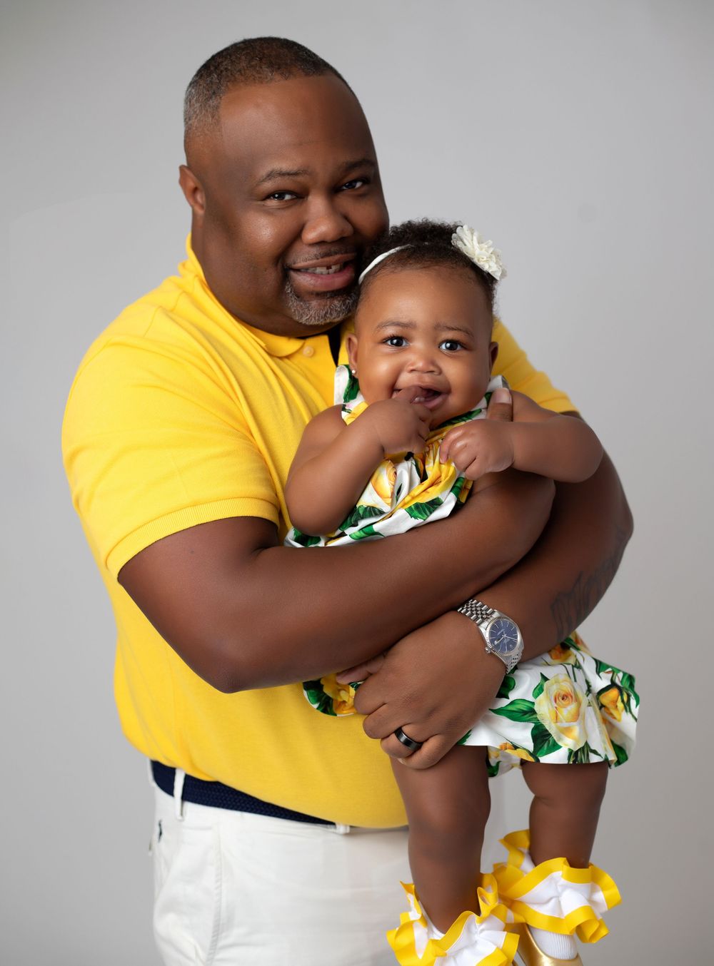 A smiling man in a yellow polo shirt holds a happy baby girl in a floral yellow dress during a studio portrait session.