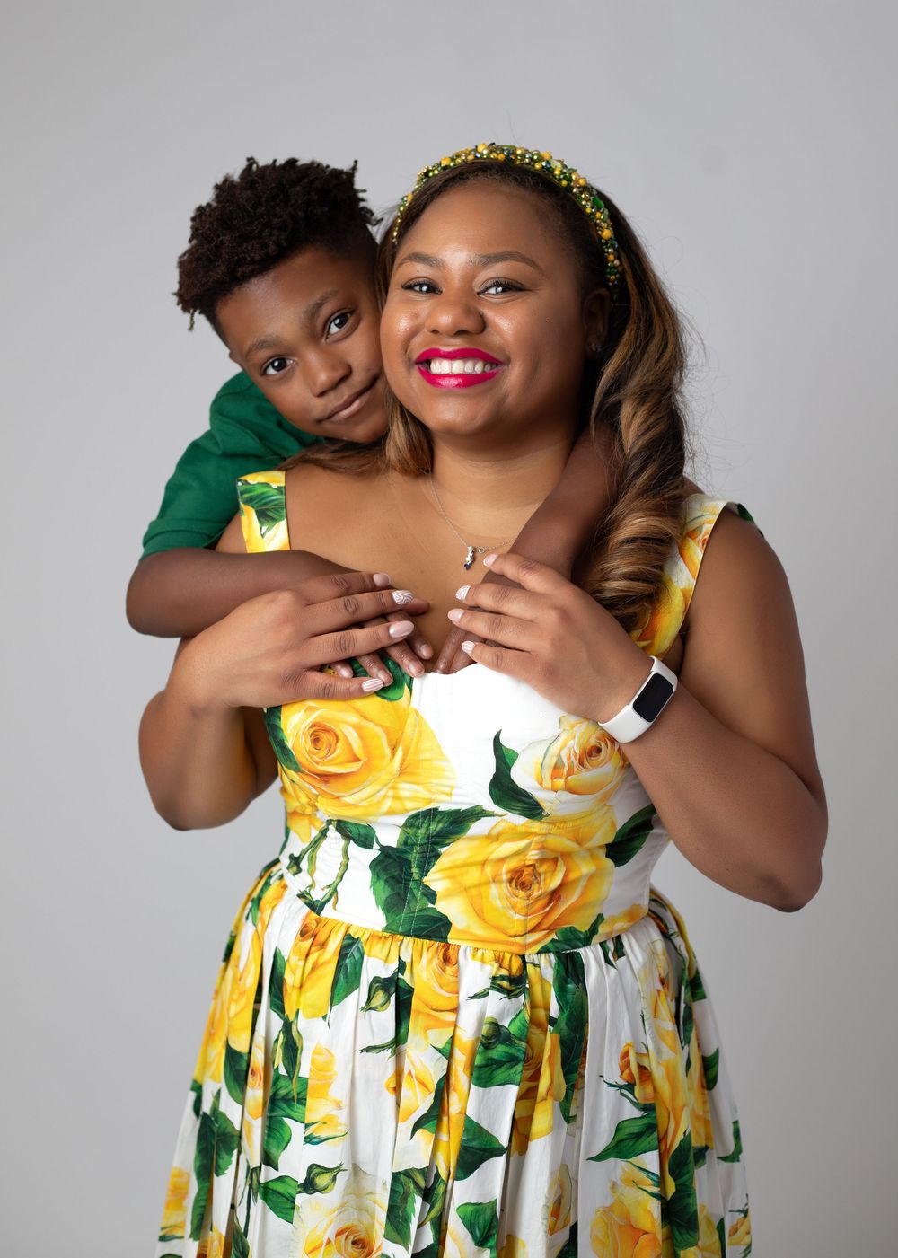 A smiling woman in a floral yellow rose dress holds a young boy in a green shirt in a studio portrait.
