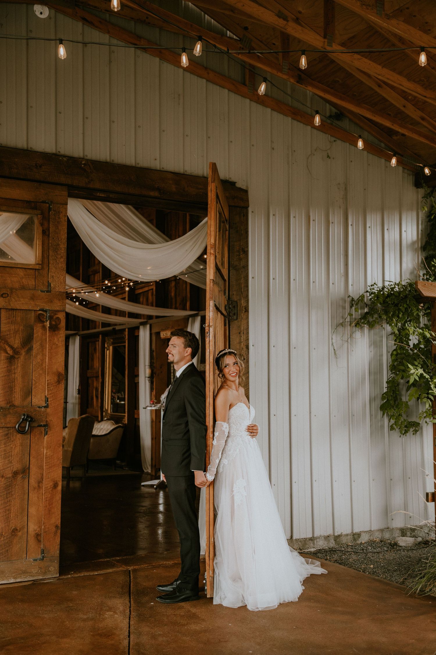 A bride and groom hold hands outside a rustic barn wedding venue adorned with string lights and draped white fabric.