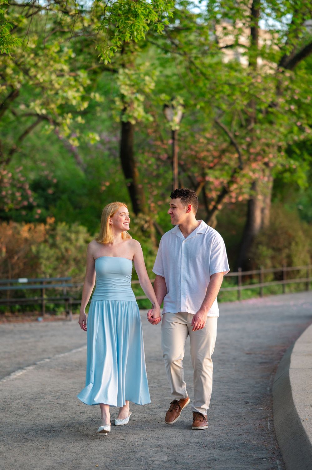 A couple holds hands and smiles while walking together on a park path surrounded by lush green trees.