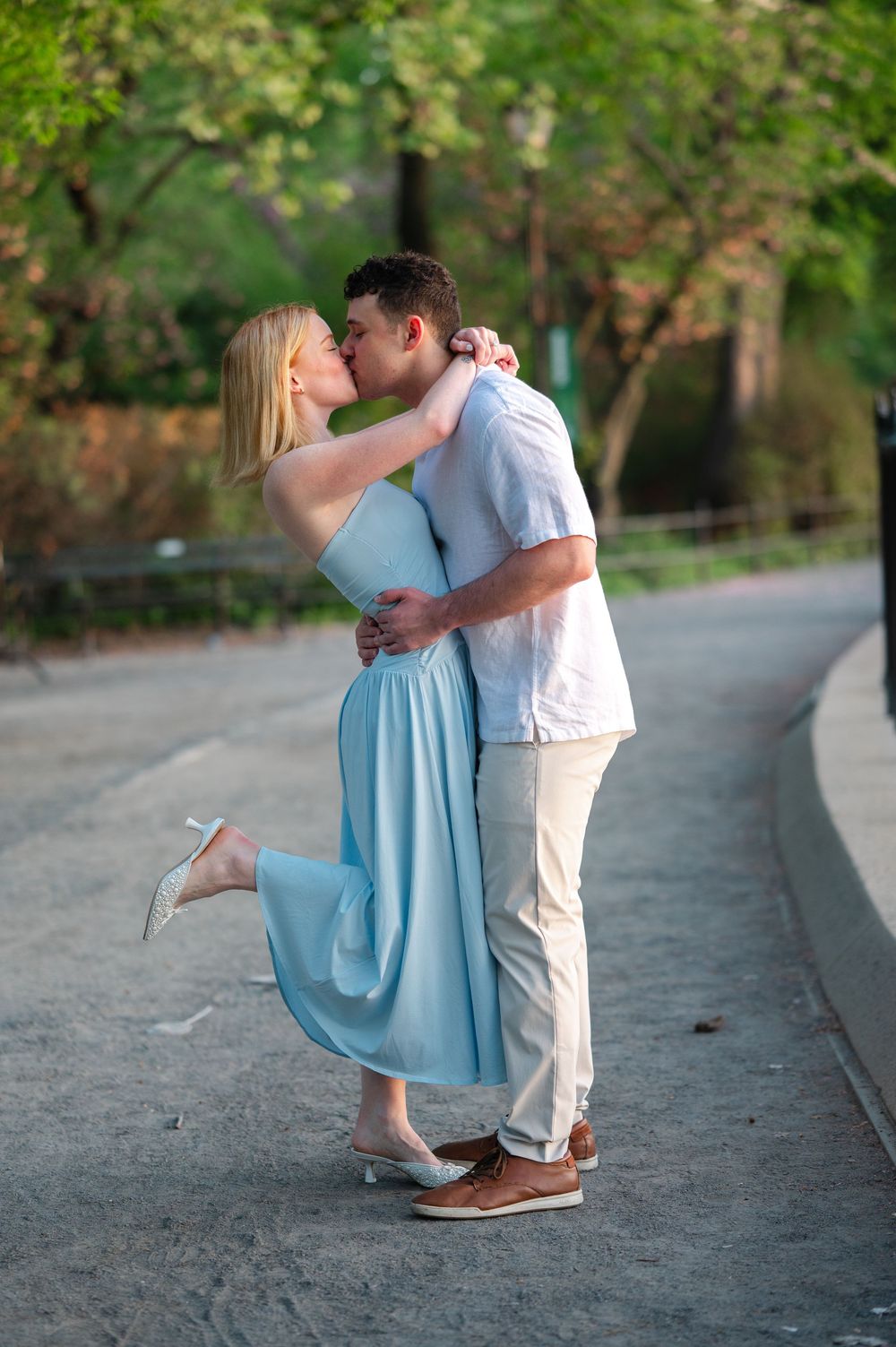 Couple sharing a romantic kiss outdoors, woman in light blue dress lifting her foot, surrounded by green trees.