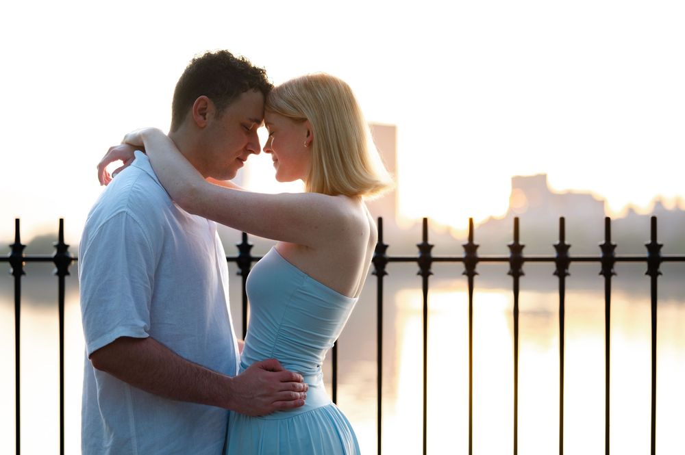 Romantic couple embracing near iron fence with city skyline and sunset in background.