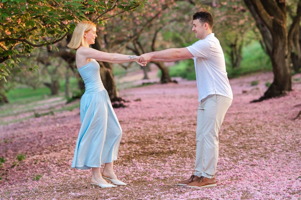 A couple dances joyfully holding hands in a pink blossom-covered park surrounded by flowering trees.