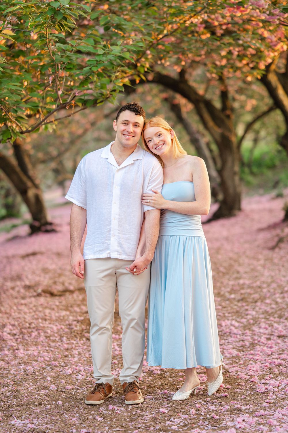 Couple smiling and holding hands under pink flowering trees with petals on ground.
