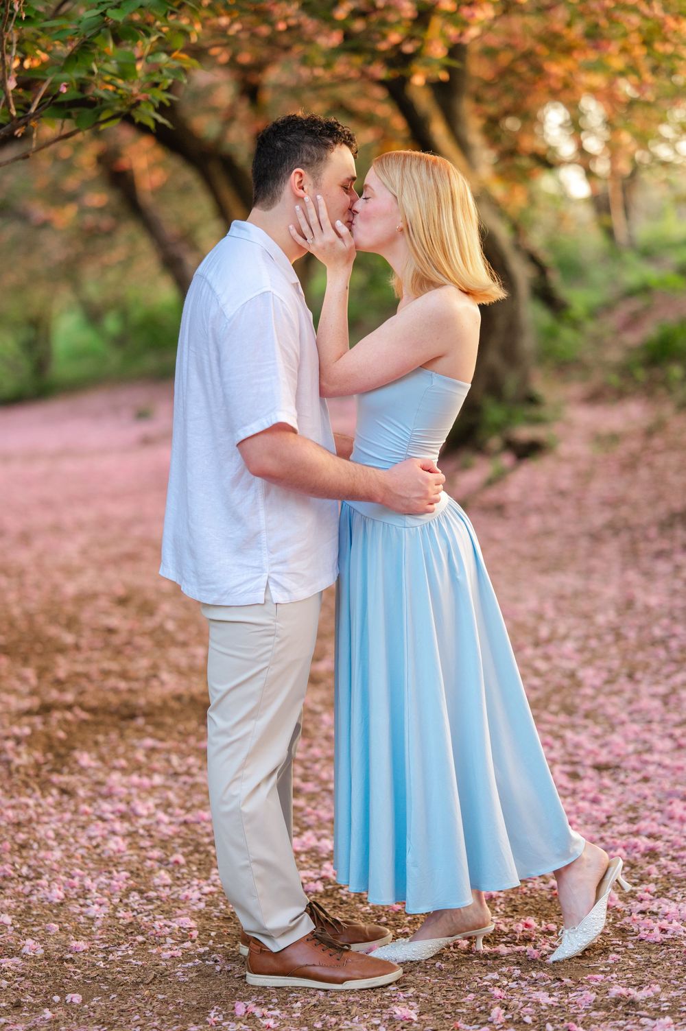 Couple sharing a romantic kiss among pink cherry blossom petals in a scenic park setting.