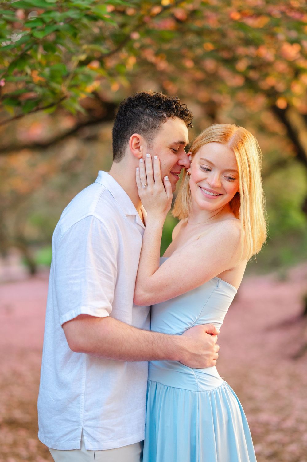 A couple embraces outdoors among autumn trees, man kissing woman's cheek as she smiles in a light blue dress.