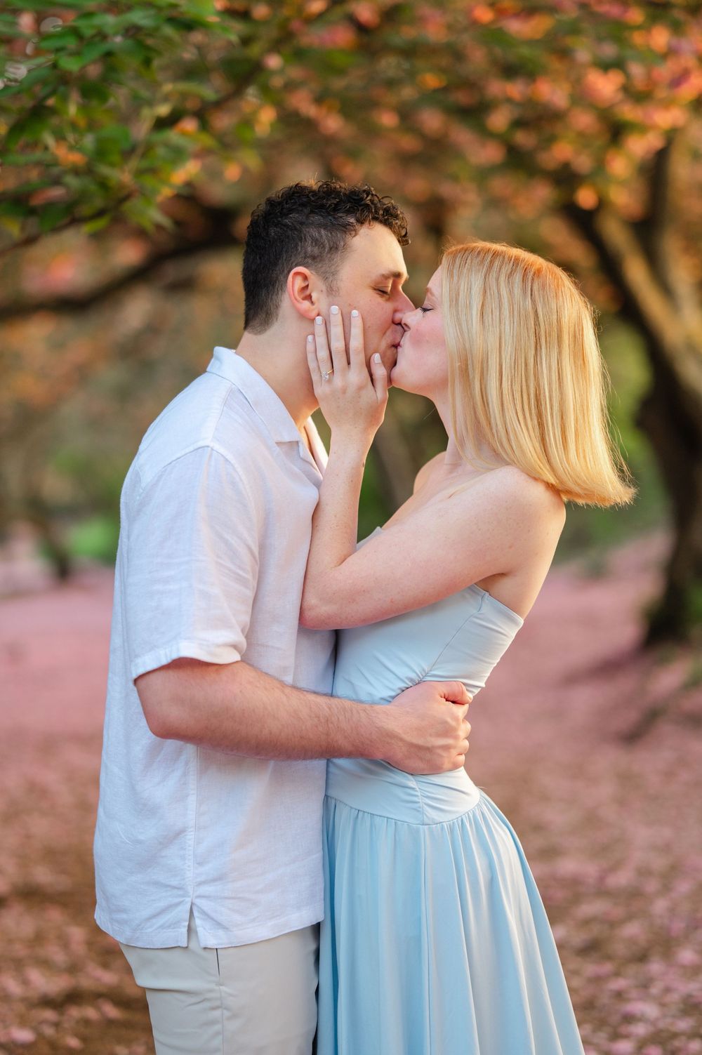 Couple sharing a romantic kiss under cherry blossom trees, woman in light blue dress touching mans face.