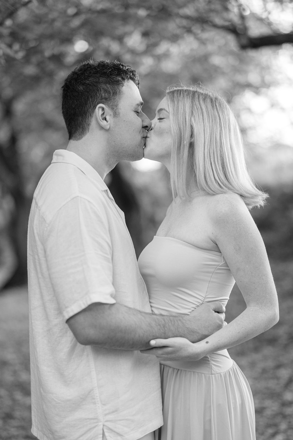 Black and white photo of a couple sharing a romantic kiss outdoors, man in white shirt embracing woman in elegant dress.