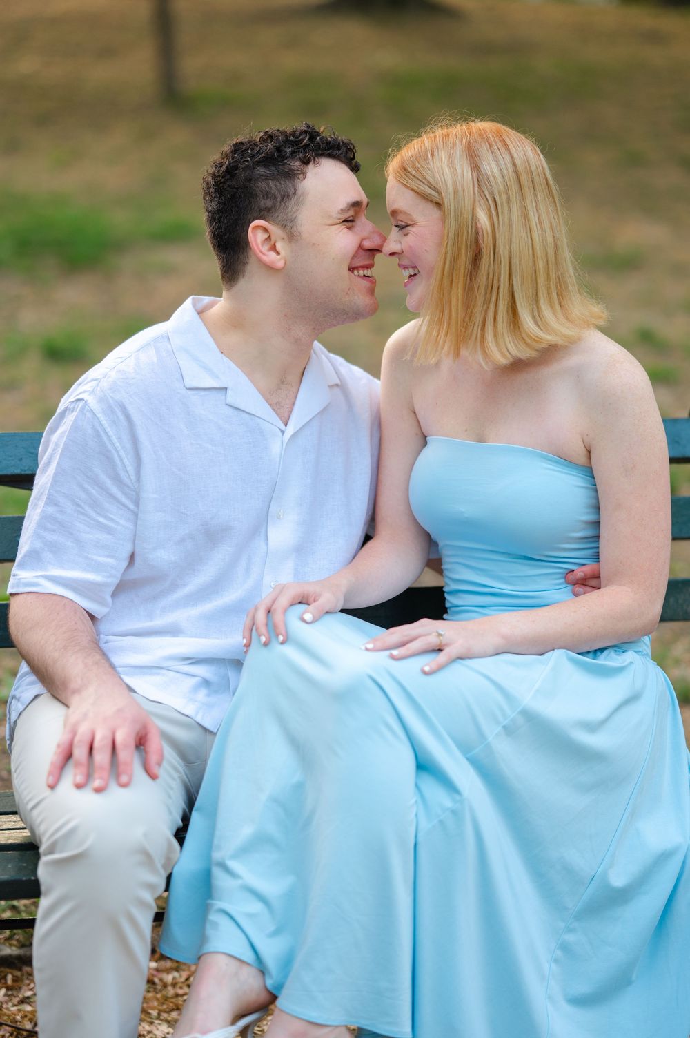 A couple in blue and white outfits sit closely on a park bench, smiling at each other in a romantic outdoor setting.