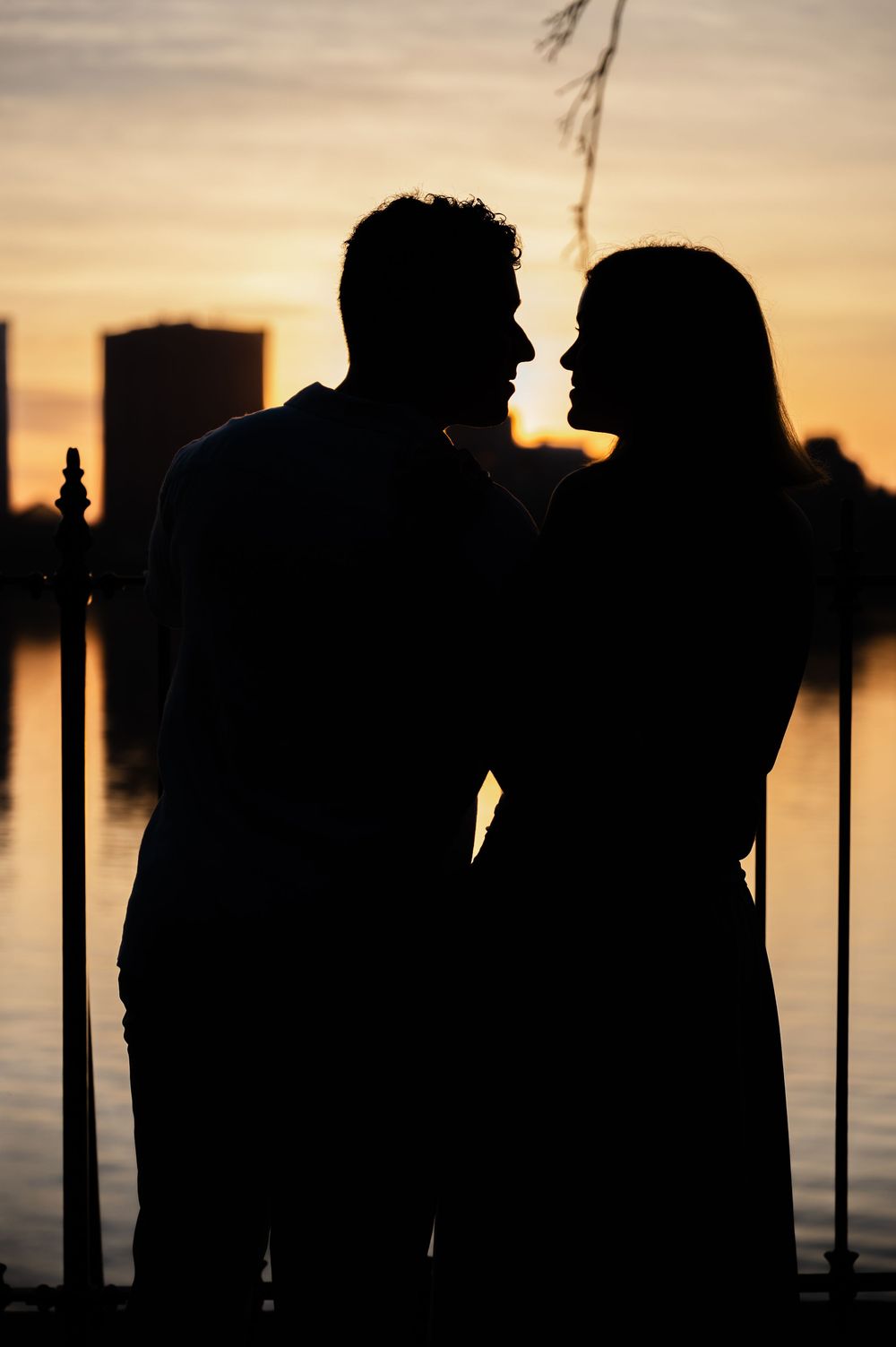 Romantic silhouette of a couple facing each other at sunset by a waterfront with a golden sky backdrop.