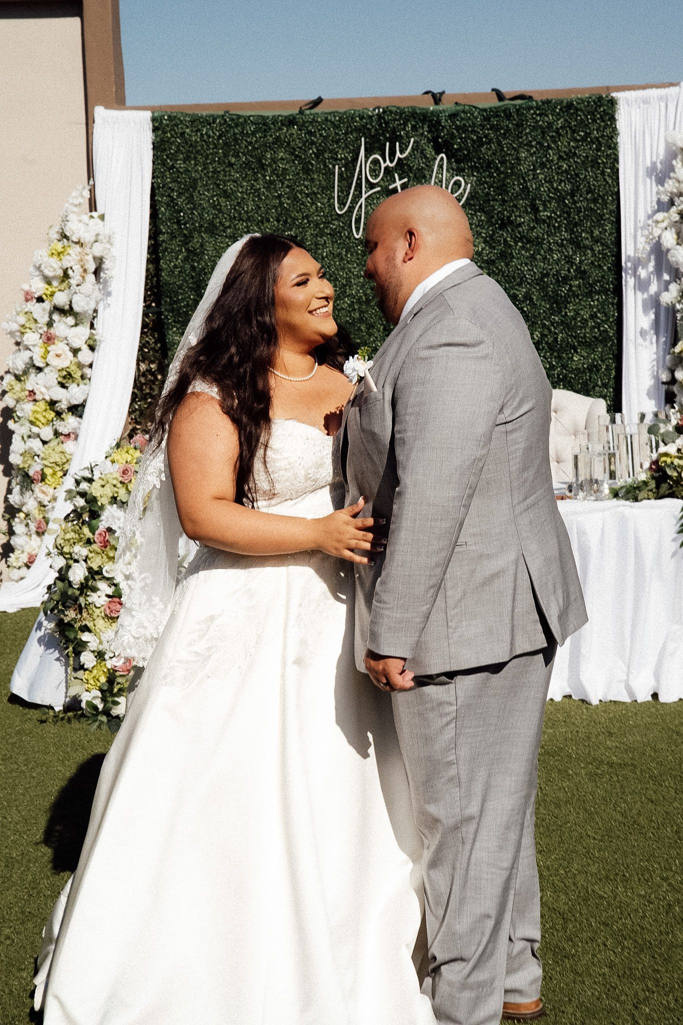 Bride in white gown and groom in gray suit share a romantic moment at an outdoor wedding ceremony with floral backdrop.