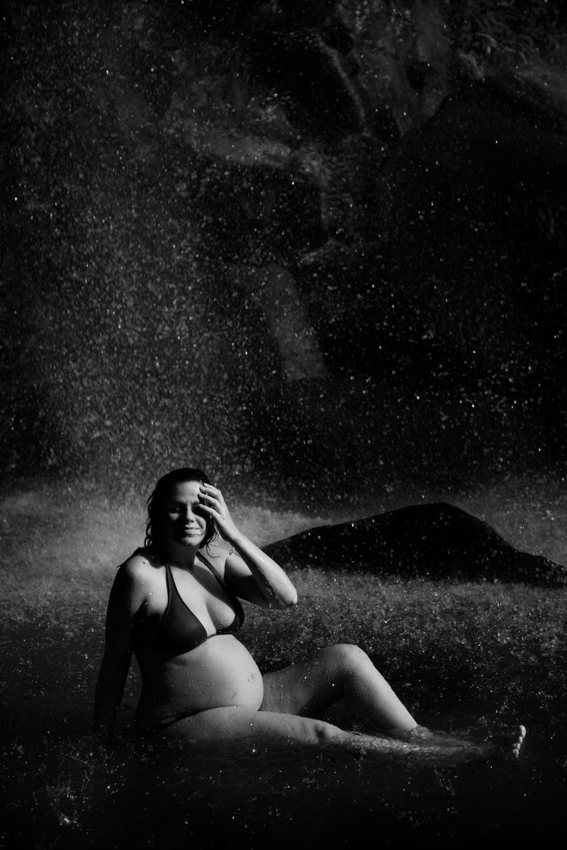 Black and white photo of a pregnant woman in a bikini sitting under a waterfall spray at night.