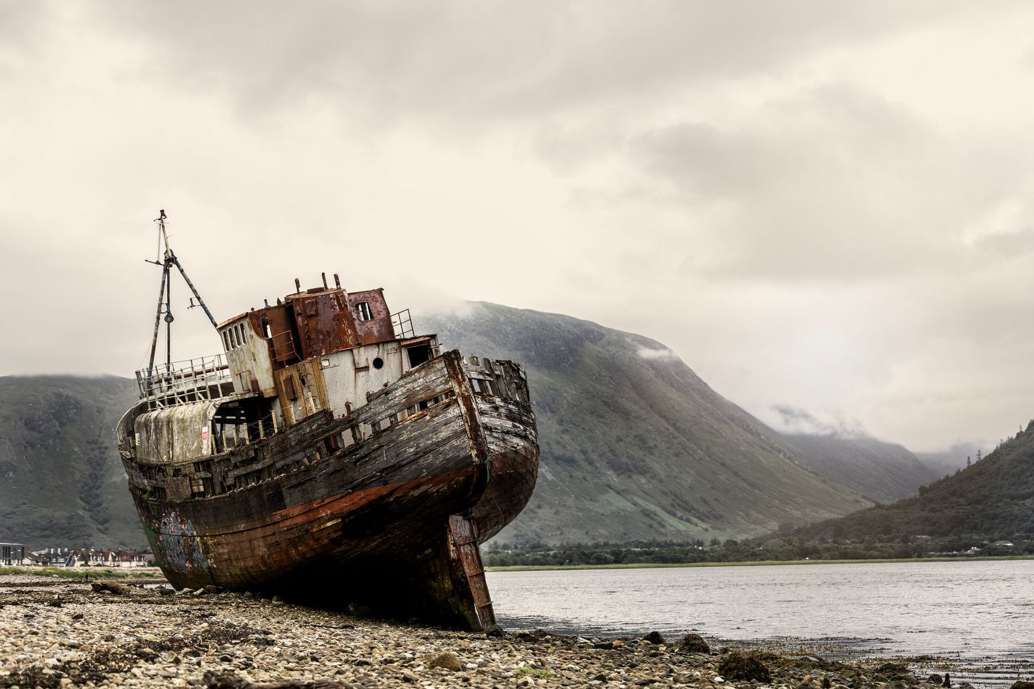 Rusty abandoned shipwreck stranded on rocky shore with misty mountains and overcast sky in Scottish Highlands.