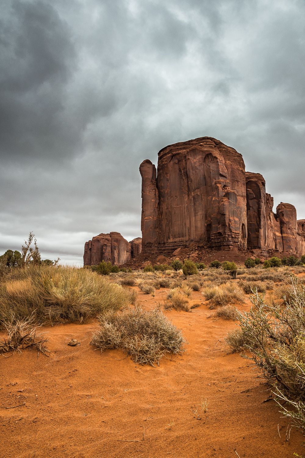 Red sandstone buttes rise above desert scrubland under dramatic stormy skies in Monument Valley, Arizona.