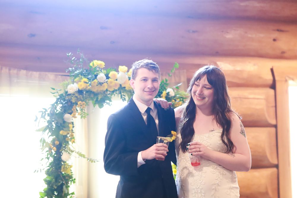 A smiling bride and groom stand before a floral arch with yellow roses at their rustic log cabin wedding ceremony.