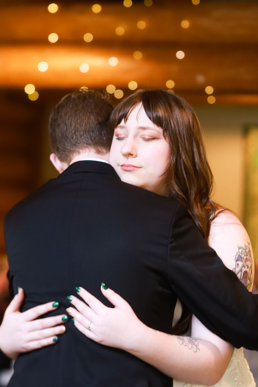 Bride with tattoos and green nails embraces groom in black suit during first dance at wedding reception.
