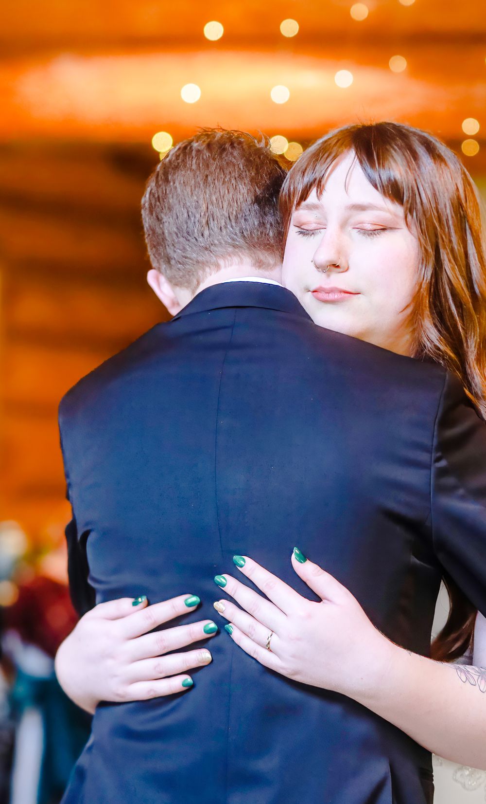 Bride with green nails and wedding ring embraces groom in navy suit during first dance at rustic venue.