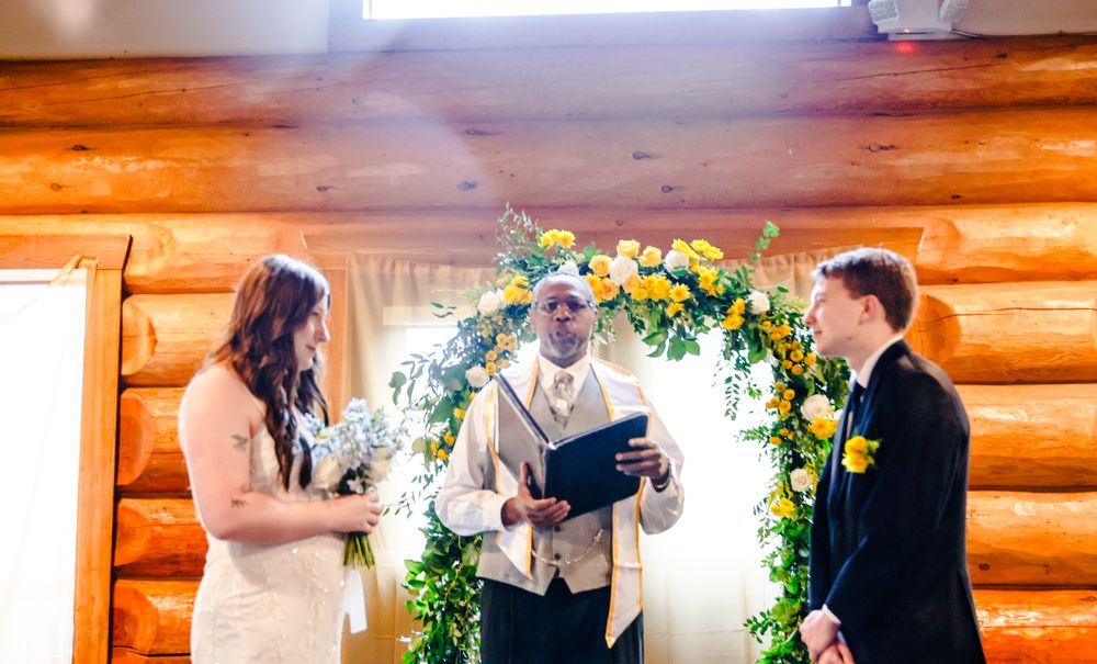 Couple exchanging vows at rustic log cabin wedding ceremony with floral arch of yellow and green flowers.