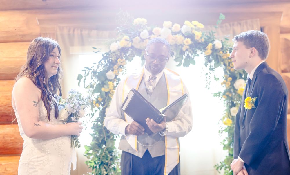 A couple exchanges wedding vows before an officiant beneath a floral arch with yellow and white flowers in a log cabin.