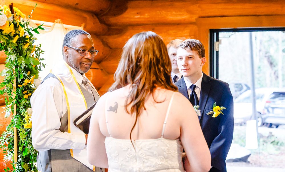 Wedding officiant smiles as bride and groom exchange vows in a cozy log cabin ceremony.