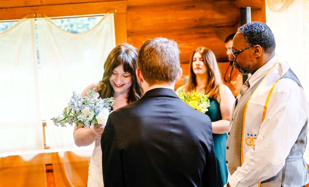 Bride and groom exchange vows at rustic wood cabin wedding ceremony with officiant and bridesmaid.