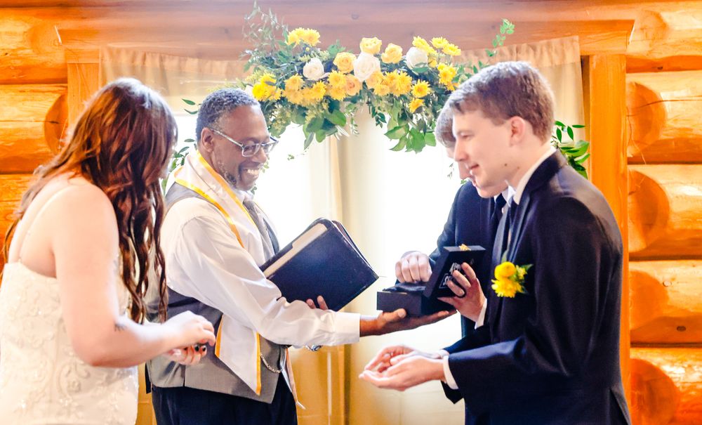 Groom exchanges rings with partner during wedding ceremony officiated by minister in rustic log cabin venue.