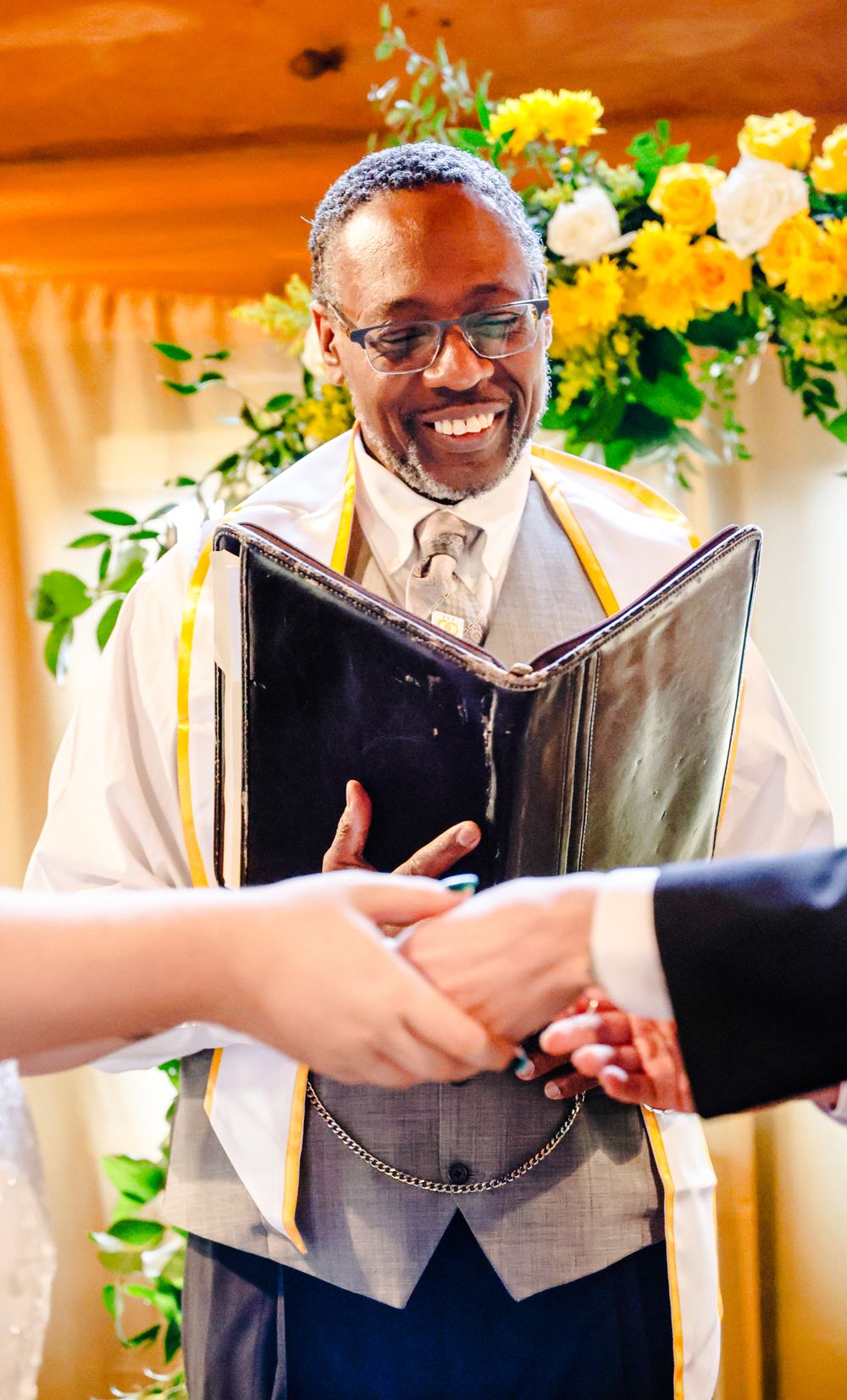 Smiling officiant holding ceremony book joins hands with couple at wedding altar with yellow floral backdrop.