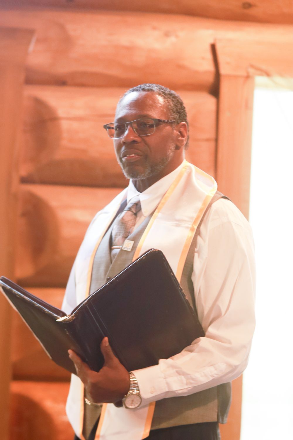 Officiant in vest and stole holds black binder during wedding ceremony in rustic log cabin venue.