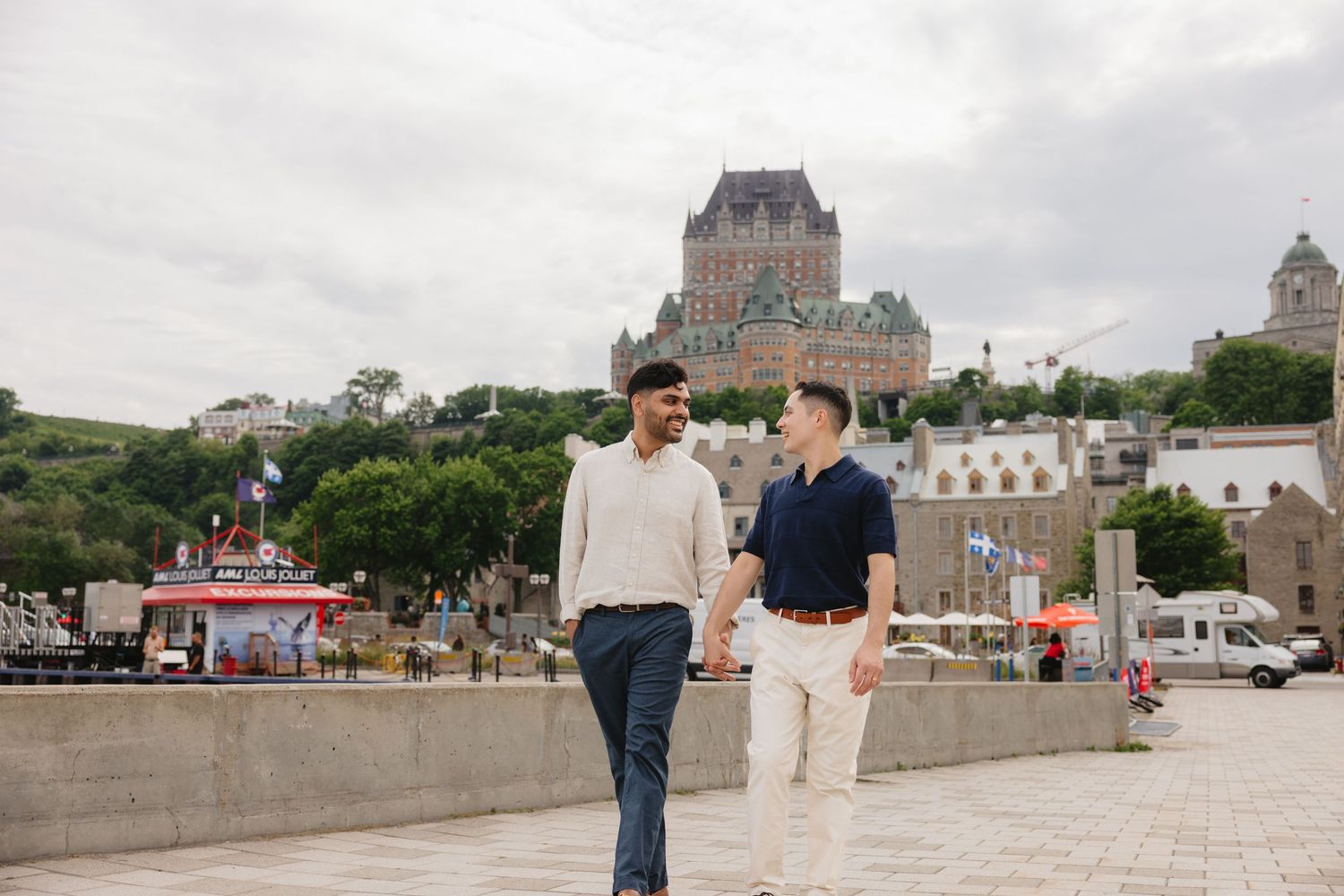 Two men walk hand in hand near Château Frontenac in Old Quebec City on a cloudy day.