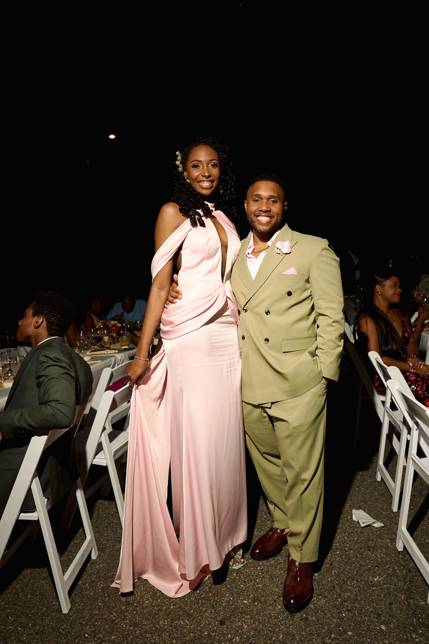 Elegant couple posing at formal outdoor gala, woman in pink gown, man in tan suit, seated guests in background.