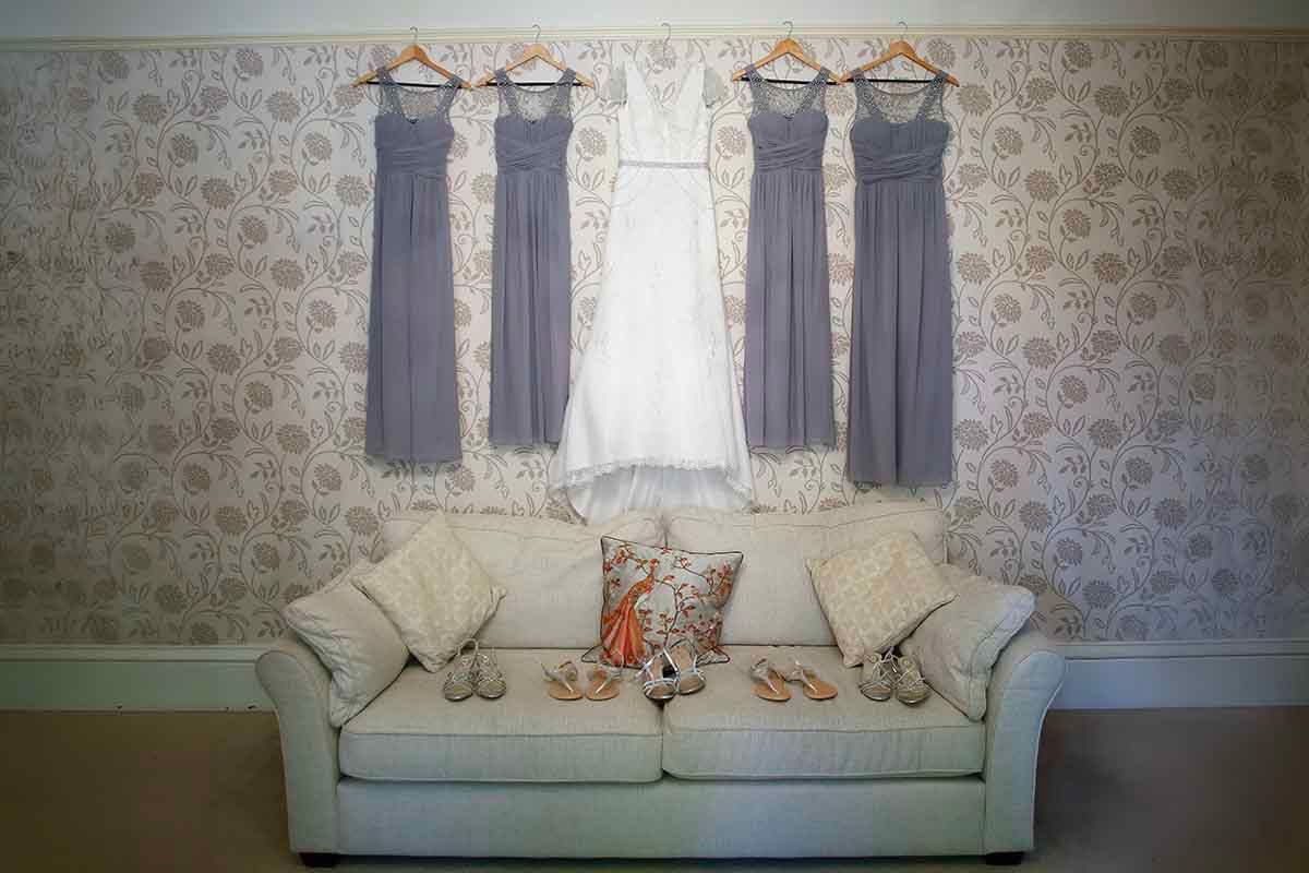 Wedding gown and lavender bridesmaid dresses hanging on wall above a white sofa with shoes and accessories displayed.