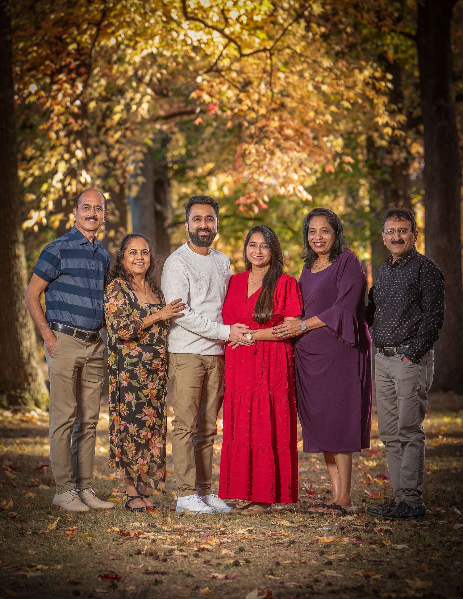 Family of six posing together in autumn park with colorful fall foliage, celebrating a pregnancy announcement outdoors.