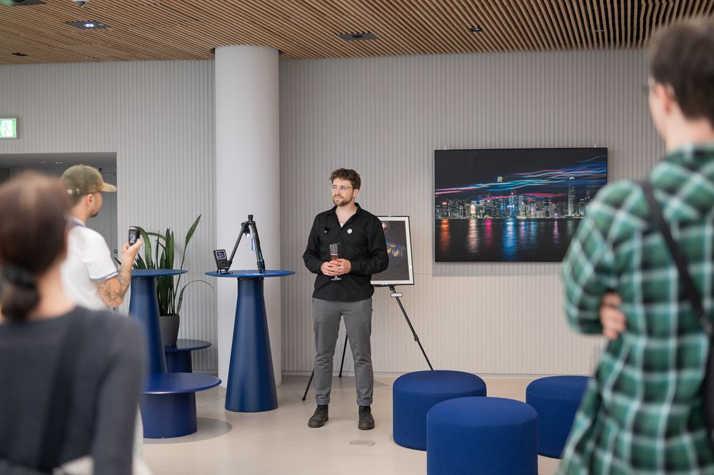 A man presents in a modern office lobby with blue furniture, a cityscape photo, and attentive audience members.