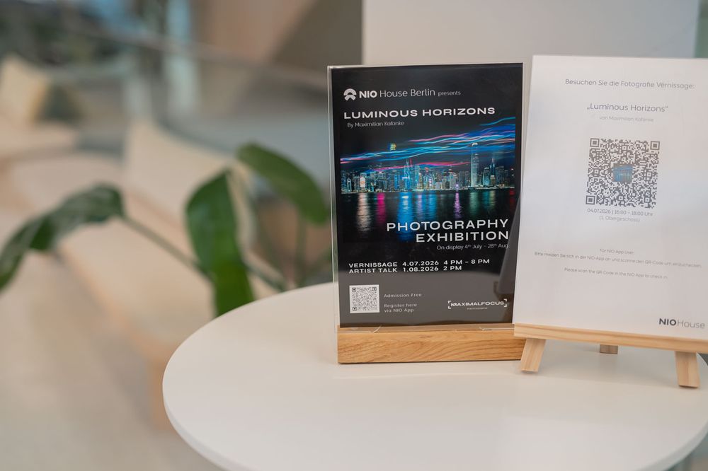 A photography exhibition display stand featuring the Luminous Horizons show poster with a QR code on a white table.