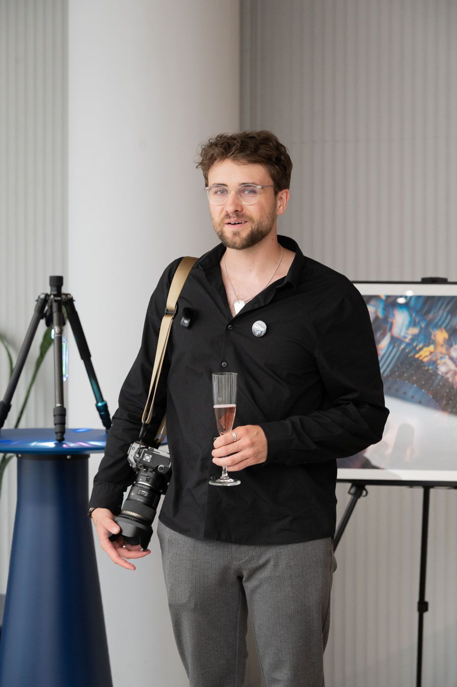 A man in a black shirt holds a champagne glass and camera at a photography exhibition event.