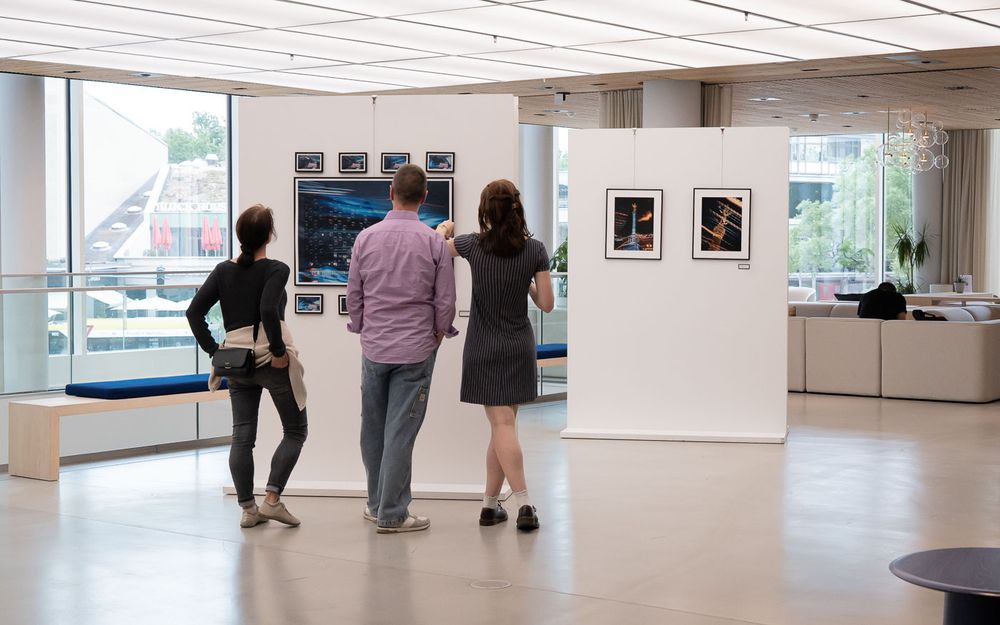 Three visitors viewing framed artwork and photography prints on display in a modern gallery exhibition space.